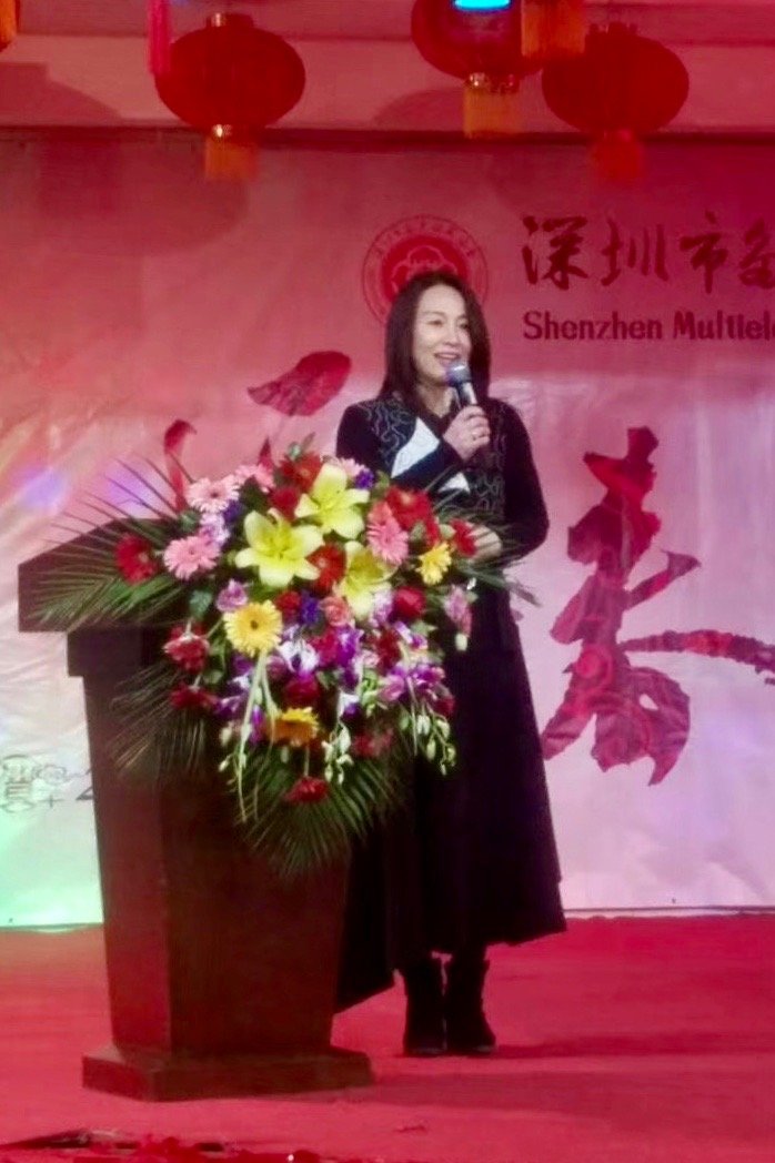 Kehan Li speaking at a lectern with a microphone during the China Collector Association event in Shenzhen, with red lanterns and floral arrangements behind her.